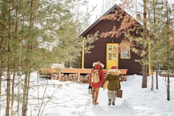 A small wooden cabin in a forest setting with two people walking toward it. 